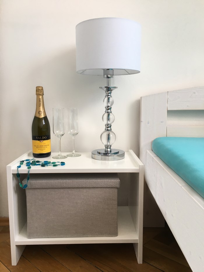 Der kleine Tisch fertig lackiert neben dem Bett. Darauf steht eine Tischleuchte und eine Flasche Sekt mit zwei Gläsern.